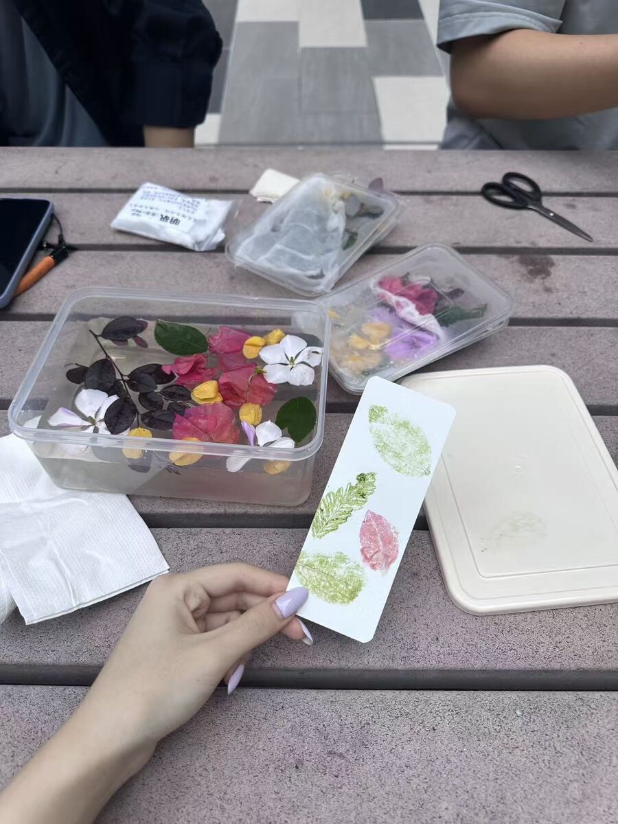 桌上摆着一盒紫叶酢浆草和各色小花，手里拿着刚敲好的书签